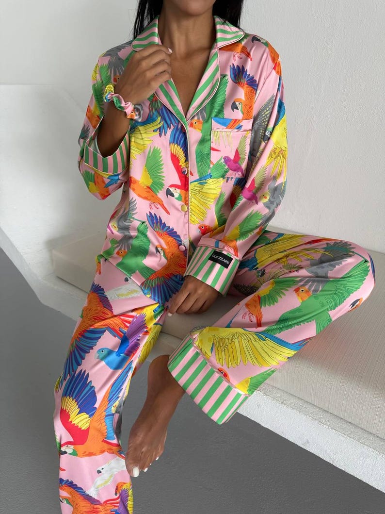 May include: Pink pajama set with a vibrant parrot print in shades of green, yellow, orange, and blue. The long-sleeved top and pants feature green and white striped accents. The set is made of a silky material.