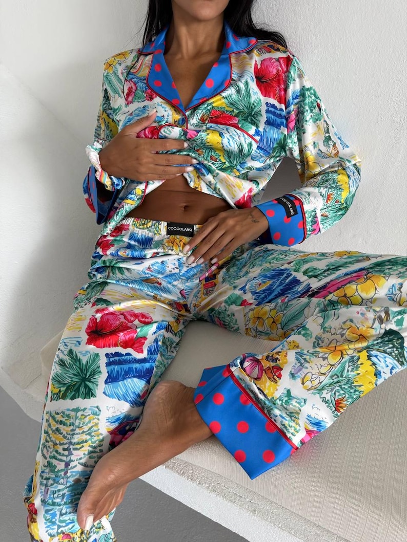 May include: A two-piece pajama set featuring a vibrant floral print in red, yellow, and green on a white background. The set includes a button-down shirt with blue trim and red polka dots, and matching pants. The pajama set is made of a silky material.