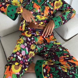 May include: A floral pajama set featuring a dark purple background with sunflowers, squirrels, and other colorful flowers. The long-sleeved top has green trim and polka dot cuffs. The matching pants complete the set, perfect for lounging.
