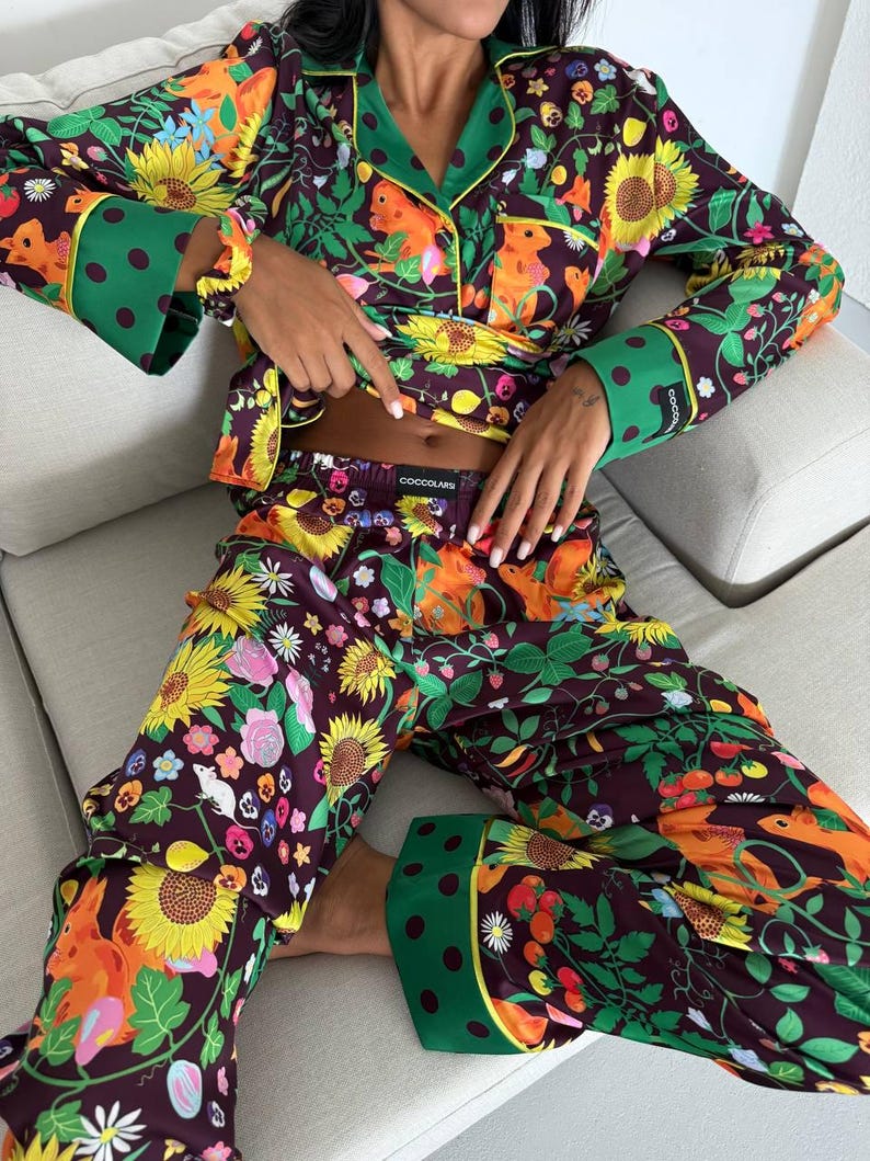 May include: A floral pajama set featuring a dark purple background with sunflowers, squirrels, and other colorful flowers. The long-sleeved top has green trim and polka dot cuffs. The matching pants complete the set, perfect for lounging.