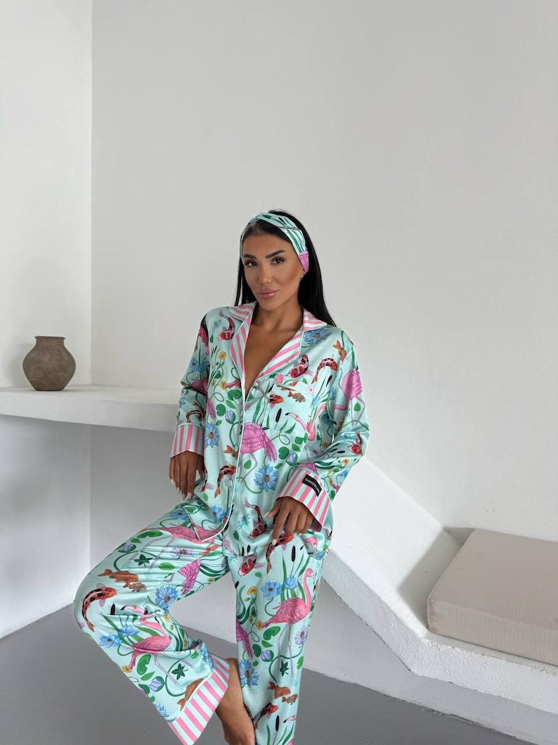 May include: A light blue pajama set with a flamingo and koi fish pattern. The set includes a long-sleeved button-down shirt, pants, and a matching headband. Pink and white striped trim accents the collar, cuffs, and pants hem.