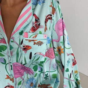 May include: Light blue pajama top with a vibrant print of pink flamingos, koi fish, and aquatic plants. The top features a collared neckline, a breast pocket, and pink and white striped trim. The sleeve cuff has the word "COCCO" printed on it.