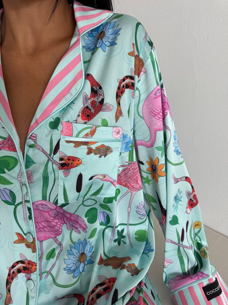 May include: Light blue pajama top with a vibrant print of pink flamingos, koi fish, and aquatic plants. The top features a collared neckline, a breast pocket, and pink and white striped trim. The sleeve cuff has the word "COCCO" printed on it.