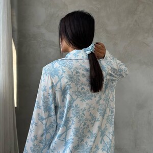 Pode incluir: Conjunto de pijama branco e azul claro com um padr&atilde;o floral e de p&aacute;ssaros. A parte superior de manga comprida tem uma frente abotoada e uma gola cl&aacute;ssica. Uma scrunchie azul claro prende o cabelo escuro num rabo de cavalo.