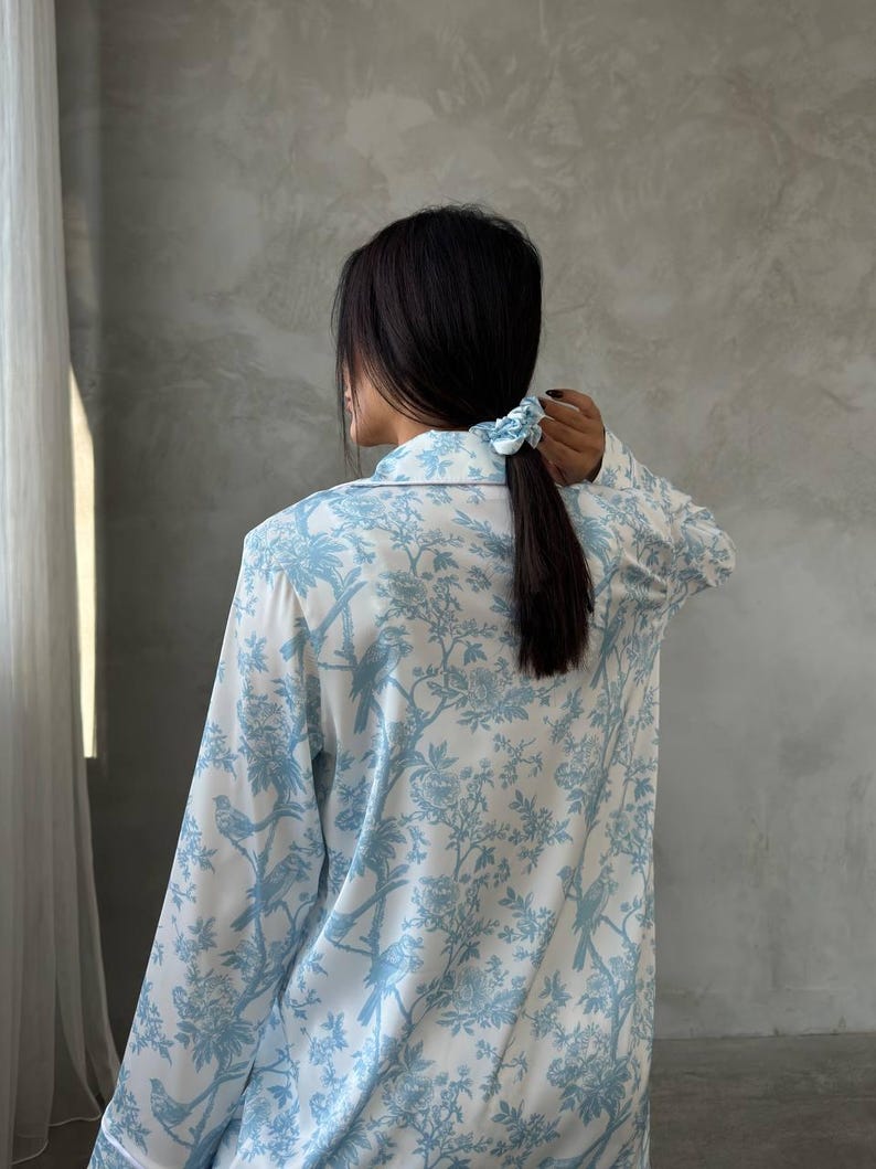 Pode incluir: Conjunto de pijama branco e azul claro com um padr&atilde;o floral e de p&aacute;ssaros. A parte superior de manga comprida tem uma frente abotoada e uma gola cl&aacute;ssica. Uma scrunchie azul claro prende o cabelo escuro num rabo de cavalo.