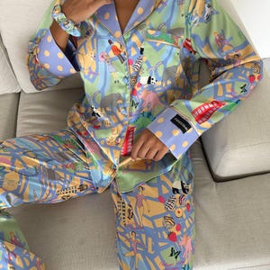 May include: A two-piece pajama set in a light blue, green, and yellow color scheme, featuring a whimsical London-themed print. The set includes a button-down shirt and pants, both adorned with illustrations of elephants, people, and red telephone booths. The cuffs and collar are accented with a polka dot pattern.