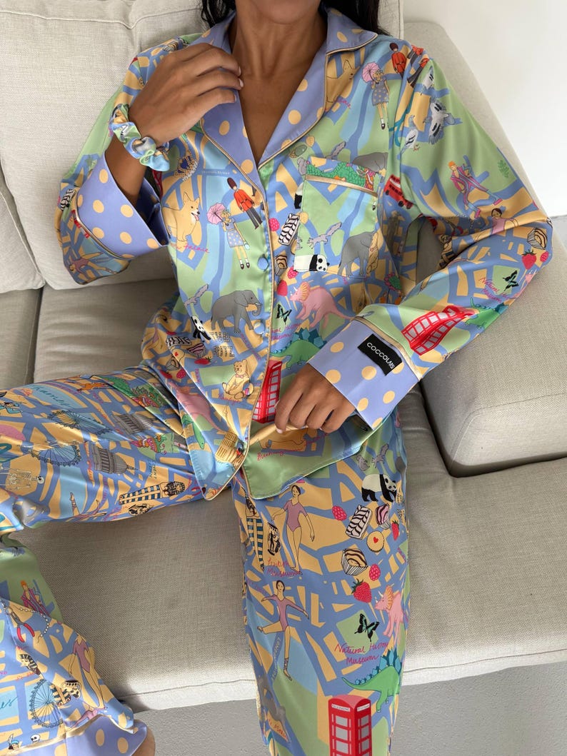 May include: A two-piece pajama set in a light blue, green, and yellow color scheme, featuring a whimsical London-themed print. The set includes a button-down shirt and pants, both adorned with illustrations of elephants, people, and red telephone booths. The cuffs and collar are accented with a polka dot pattern.