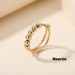 Puede incluir: Un anillo dorado con un diseño de doble banda. La banda superior presenta seis pequeñas cuentas esféricas espaciadas uniformemente. El anillo está sobre un fondo neutro, con el nombre de la marca "Nooréa" visible en la esquina inferior derecha.