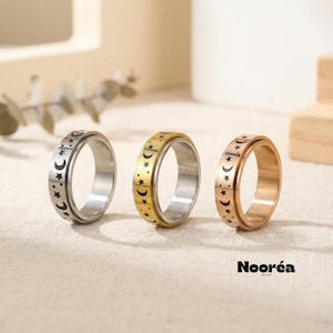 Op de afbeelding: Drie roestvrijstalen spinnerringen in zilver, goud en roségoud. Elke ring heeft een band met een herhalend patroon van halve manen en sterren. De ringen worden gepresenteerd op een neutrale ondergrond.