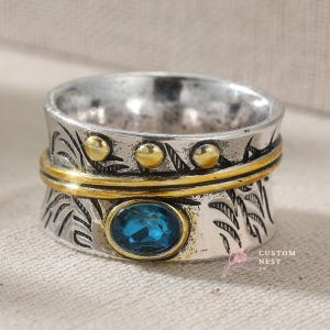 May include: A silver-tone ring with a gold band and accents. The ring features a blue gemstone and decorative leaf patterns. Three gold-colored studs are set on the top band. The ring is a unique piece of jewelry.