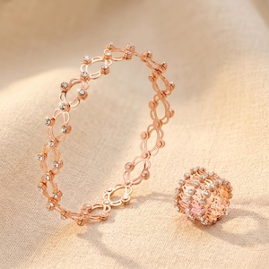 Puede incluir: Juego de joyas en tono oro rosa que incluye un brazalete y un anillo. El brazalete tiene un diseño calado con una serie de eslabones interconectados en forma de flor, cada uno adornado con pequeños cristales transparentes. El anillo refleja el diseño del brazalete.