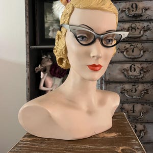 May include: A vintage mannequin head with blonde hair and red lipstick, wearing cat-eye glasses with black frames and white accents. The mannequin is displayed on a wooden surface, with a cabinet in the background.