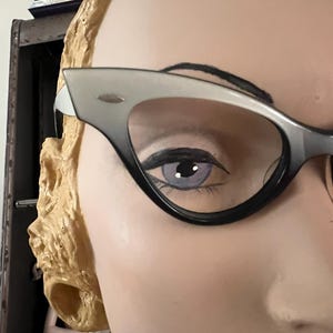 May include: Close-up of a mannequin's face wearing cat-eye glasses. The glasses have a black frame that fades to gray at the top. The mannequin has a painted eye with a light purple iris and black eyeliner. The mannequin's hair is styled in blonde curls.