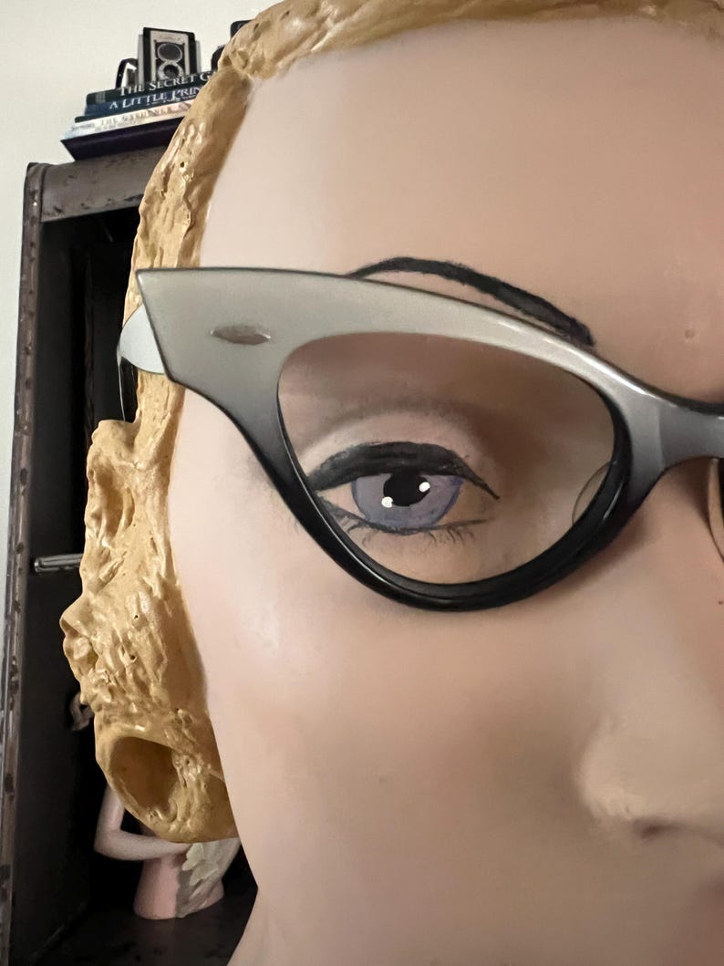 May include: Close-up of a mannequin's face wearing cat-eye glasses. The glasses have a black frame that fades to gray at the top. The mannequin has a painted eye with a light purple iris and black eyeliner. The mannequin's hair is styled in blonde curls.