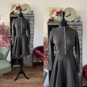 Puede incluir: Un vestido gris oscuro con mangas largas y cierre de botones en la parte delantera. El vestido tiene una falda acampanada y está en un maniquí.