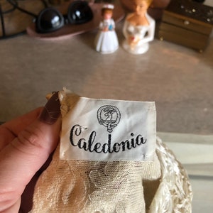 May include: A white lace fabric with a white satin flower detail. The fabric has a label that reads "Caledonia" with a crest.