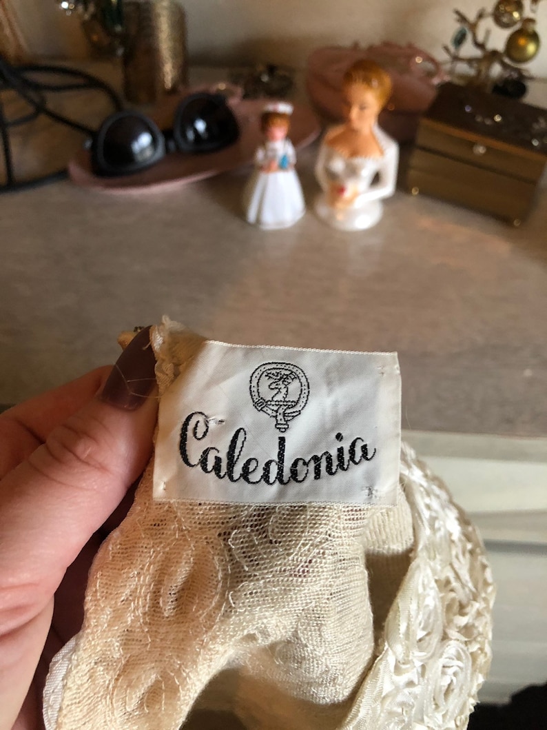 May include: A white lace fabric with a white satin flower detail. The fabric has a label that reads "Caledonia" with a crest.