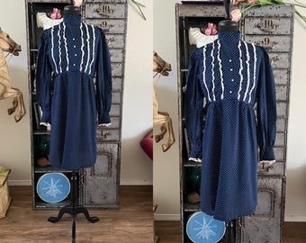 May include: A vintage navy blue dress with white polka dots and long sleeves. The dress features white ruffled trim and a high neckline. It is displayed on a black dress form in front of a vintage metal cabinet.