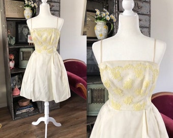 Puede incluir: Un vestido blanco vintage con bordados florales amarillos. El vestido tiene un corpiño ajustado y una falda completa. Es sin mangas y tiene un escote redondo.