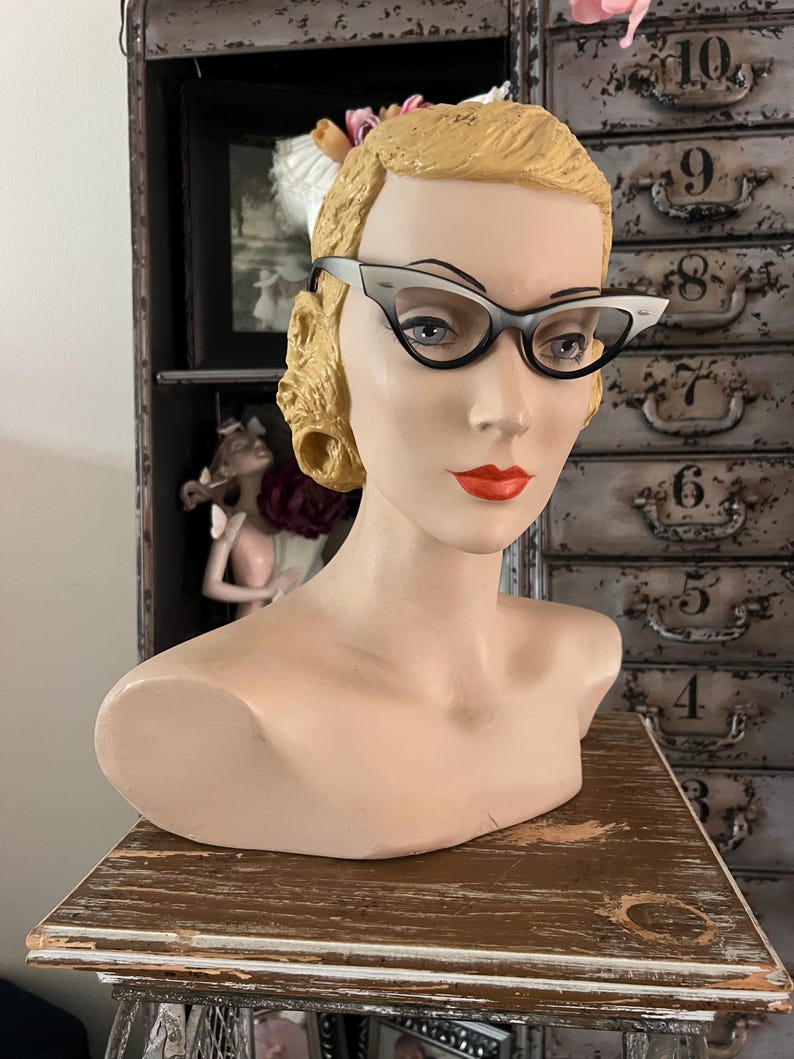 May include: A vintage mannequin head with blonde hair and red lipstick, wearing cat-eye glasses with black frames and white accents. The mannequin is displayed on a wooden surface, with a cabinet in the background.