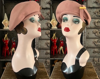 May include: Two mannequin heads wearing pink berets. The berets have a small bow and gold-colored clasp. The mannequins have red lipstick, dark eye makeup, and black tops. The background includes decorative items.