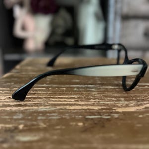 May include: Black and white cat-eye style eyeglasses. The frames have a classic design with a black upper rim and temples, transitioning to a white lower rim. The glasses are set against a textured, light brown surface.