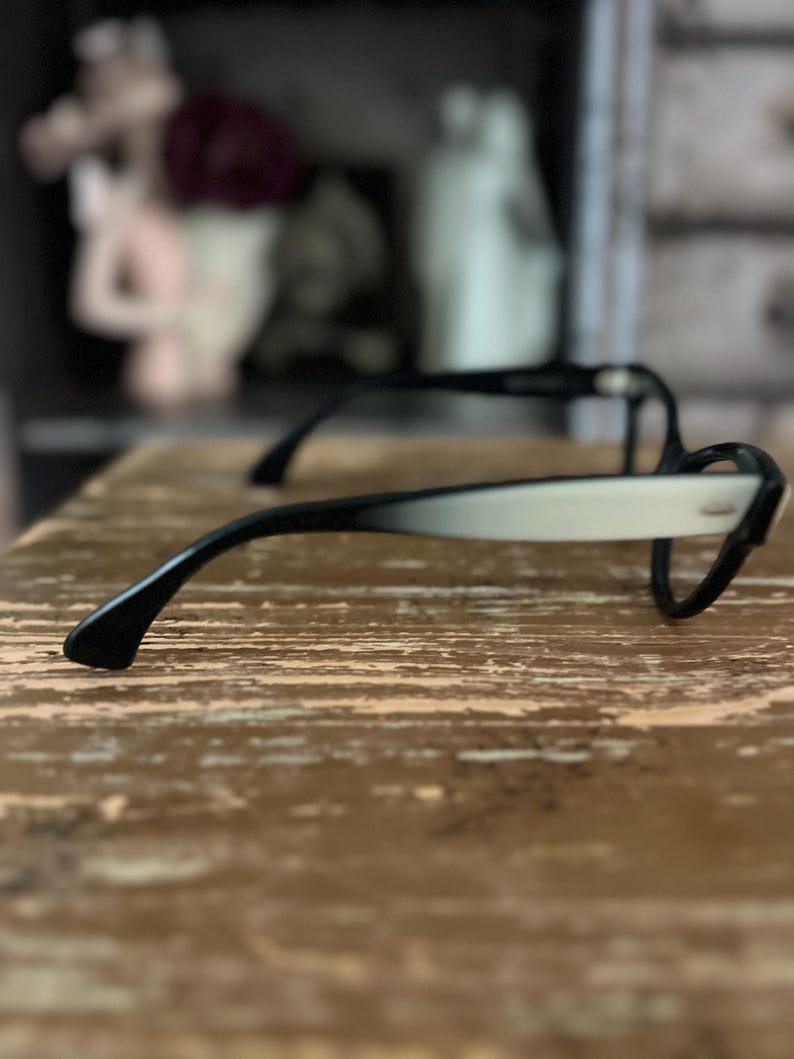 May include: Black and white cat-eye style eyeglasses. The frames have a classic design with a black upper rim and temples, transitioning to a white lower rim. The glasses are set against a textured, light brown surface.