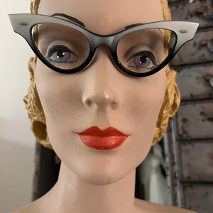 May include: Cat-eye style eyeglasses with a two-tone gray and black frame. The glasses are displayed on a mannequin head with red lipstick and a vintage hairstyle. The frames have a classic, retro design.