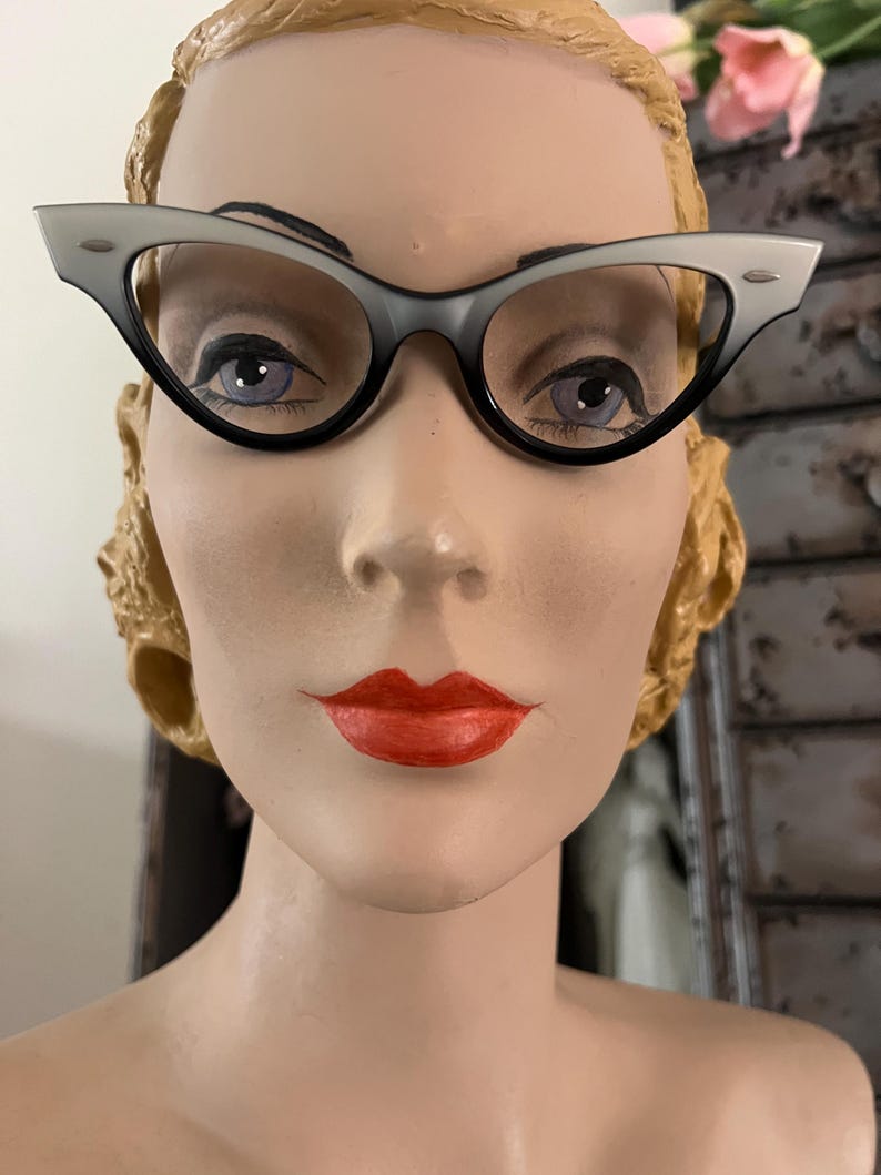 May include: Cat-eye style eyeglasses with a two-tone gray and black frame. The glasses are displayed on a mannequin head with red lipstick and a vintage hairstyle. The frames have a classic, retro design.