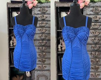 Può includere: Un costume da bagno blu reale in stile vintage con corpetto e gonna arricciati. Il costume da bagno ha spalline sottili e uno scollo a cuore. L'indumento è esposto su un manichino nero. Lo sfondo include un mobile metallico con cassetti.