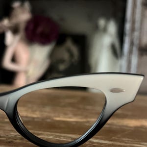 May include: Close-up of a pair of cat-eye eyeglasses. The frames are black at the bottom, transitioning to white at the top. A small silver accent is visible on the upper frame. The glasses rest on a wooden surface.