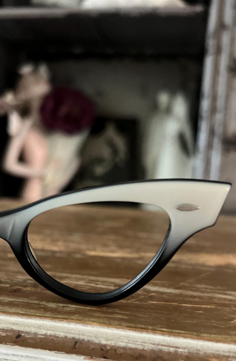 May include: Close-up of a pair of cat-eye eyeglasses. The frames are black at the bottom, transitioning to white at the top. A small silver accent is visible on the upper frame. The glasses rest on a wooden surface.