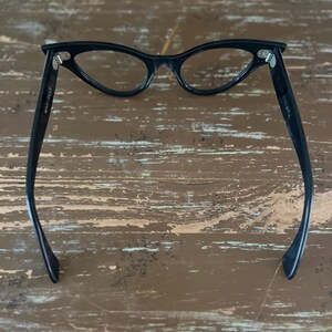 May include: Black cat-eye glasses with a classic design. The glasses have a sleek, curved frame and clear lenses. The arms of the glasses are extended and feature the text "MADE IN USA".