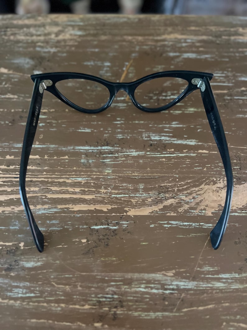 May include: Black cat-eye glasses with a classic design. The glasses have a sleek, curved frame and clear lenses. The arms of the glasses are extended and feature the text "MADE IN USA".
