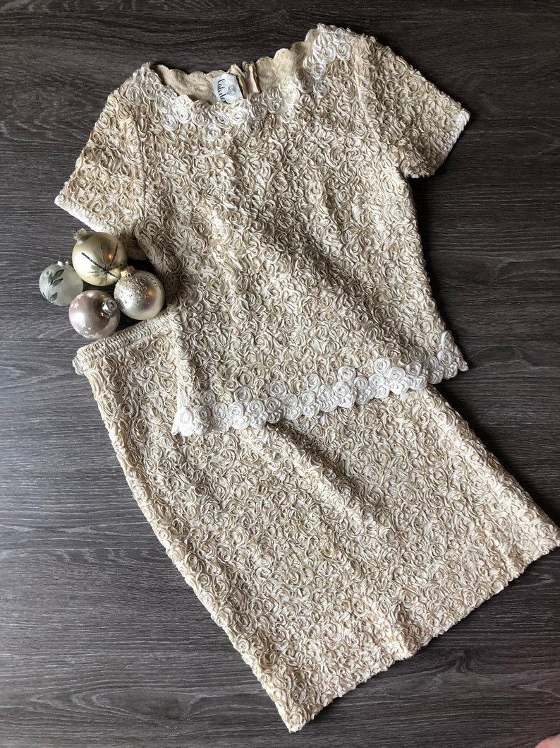 May include: A two-piece outfit in a cream color with a rose-like textured fabric. The top has short sleeves and a round neckline. The skirt is a pencil skirt.