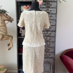 May include: A white dress with a rose pattern and a white lace trim. The dress is on a mannequin and is in front of a metal cabinet with numbered drawers.