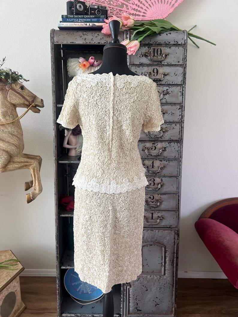 May include: A white dress with a rose pattern and a white lace trim. The dress is on a mannequin and is in front of a metal cabinet with numbered drawers.
