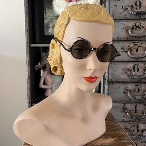 May include: A mannequin head with blonde hair and red lipstick is wearing round, tortoiseshell sunglasses. The sunglasses have a dark brown frame and lenses. The mannequin is on a wooden surface, with a metal cabinet in the background.