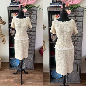 May include: A two-piece dress set in a cream color with a floral lace design. The dress has a fitted bodice and a skirt with a slight A-line shape. The dress is displayed on a mannequin.