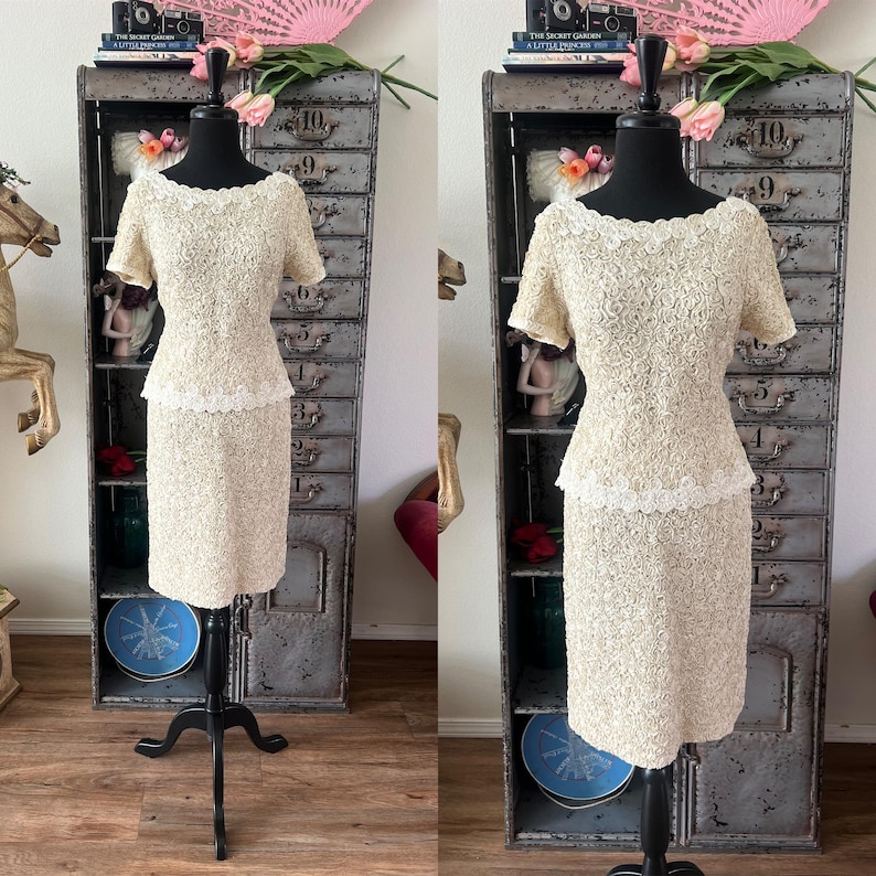 May include: A two-piece dress set in a cream color with a floral lace design. The dress has a fitted bodice and a skirt with a slight A-line shape. The dress is displayed on a mannequin.