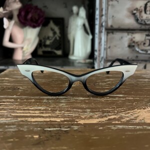 May include: Cat-eye style eyeglasses with a gradient black to white frame. The glasses have a classic retro design, with a black bridge and temples. The lenses are clear. The glasses are set on a wooden surface.