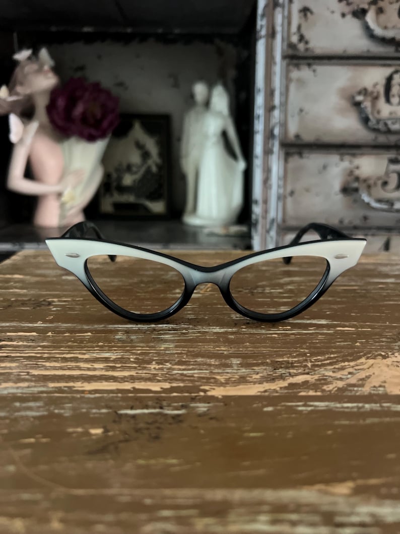 May include: Cat-eye style eyeglasses with a gradient black to white frame. The glasses have a classic retro design, with a black bridge and temples. The lenses are clear. The glasses are set on a wooden surface.