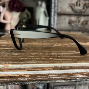 May include: Black and white eyeglasses with a retro design. The frames are black, with a gradient from white to black on the arms. The glasses are resting on a distressed wooden surface.