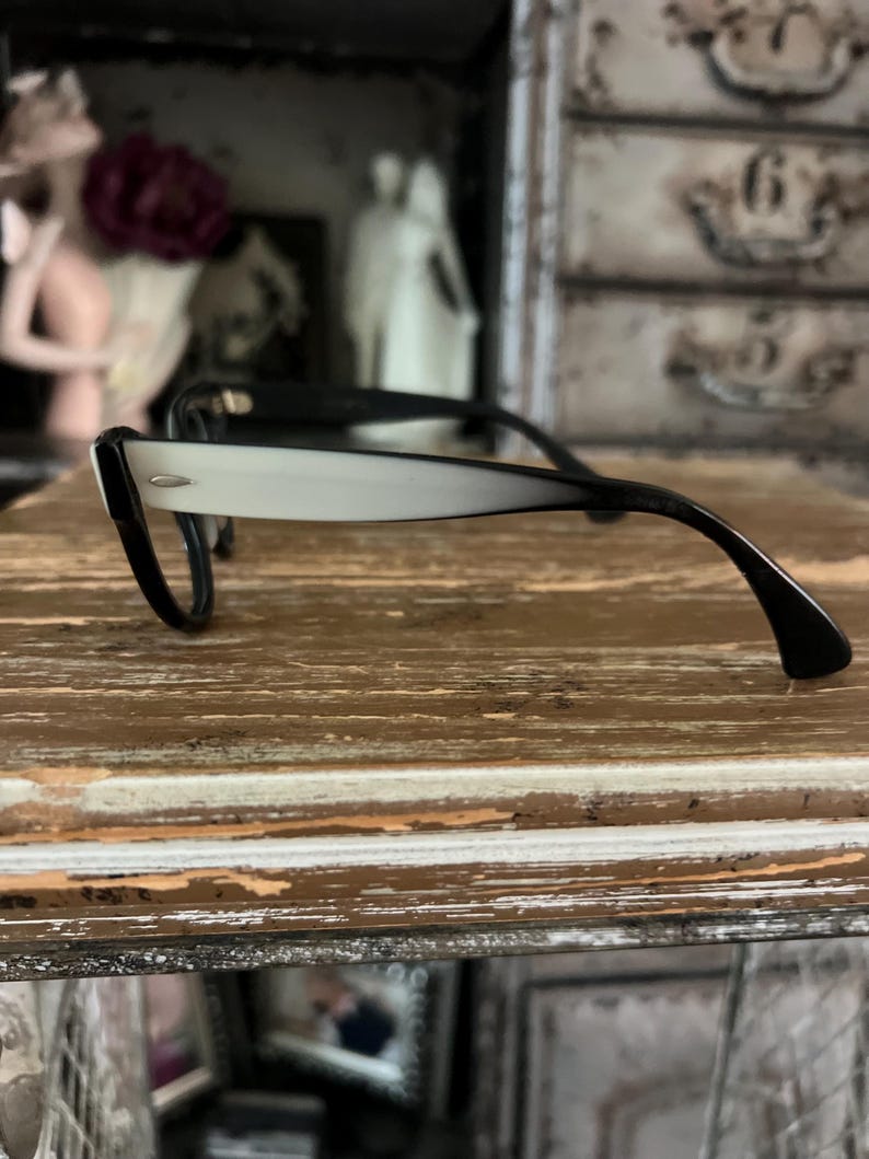 May include: Black and white eyeglasses with a retro design. The frames are black, with a gradient from white to black on the arms. The glasses are resting on a distressed wooden surface.