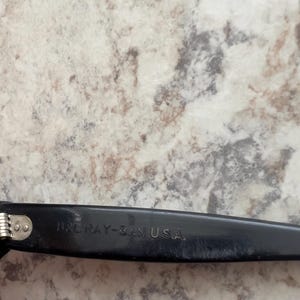 May include: Close-up of a black Ray-Ban eyeglass arm. The text "B&L RAY-BAN U.S.A." is printed in white on the arm. The hinge is silver. The background is a light-colored, speckled surface.
