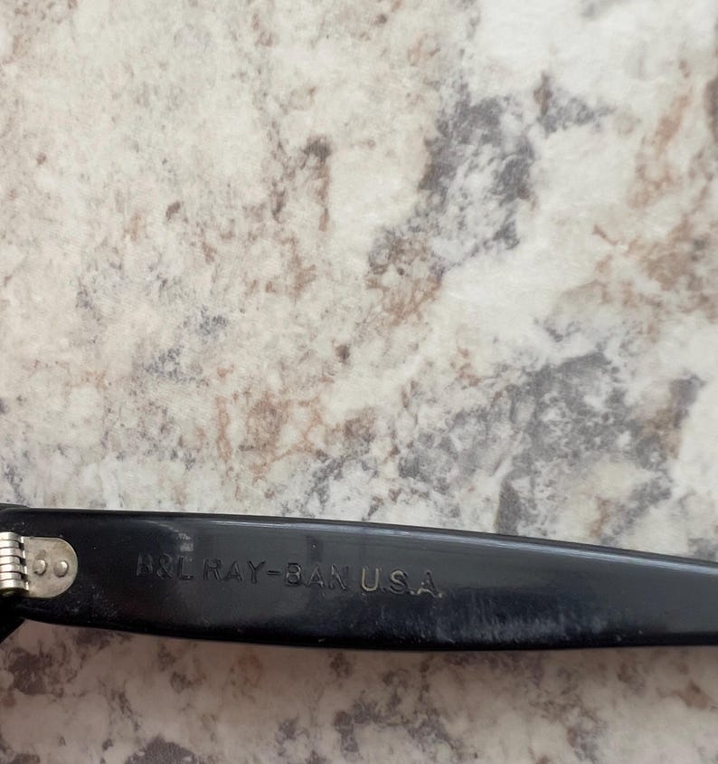 May include: Close-up of a black Ray-Ban eyeglass arm. The text "B&L RAY-BAN U.S.A." is printed in white on the arm. The hinge is silver. The background is a light-colored, speckled surface.