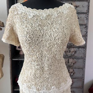 May include: A vintage ivory dress with a fitted bodice and a skirt made of a fabric with a raised rose pattern. The dress has a scoop neckline and short sleeves.