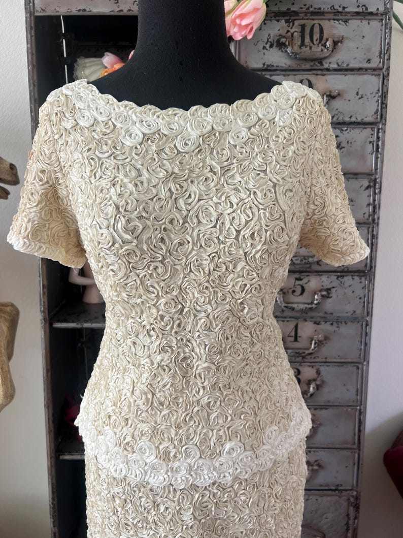 May include: A vintage ivory dress with a fitted bodice and a skirt made of a fabric with a raised rose pattern. The dress has a scoop neckline and short sleeves.