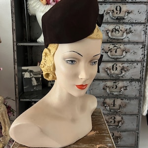 May include: A dark brown felt hat with a structured, asymmetrical crown and a small bow detail on the side. The hat is displayed on a mannequin head with blonde hair, blue eyes, and red lips, positioned against a metal cabinet with numbered drawers.