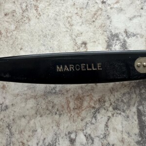 May include: Close-up of a black eyeglass frame with the name "MARCELLE" in silver lettering. The frame has a sleek, modern design. The background is a light-colored, speckled surface.